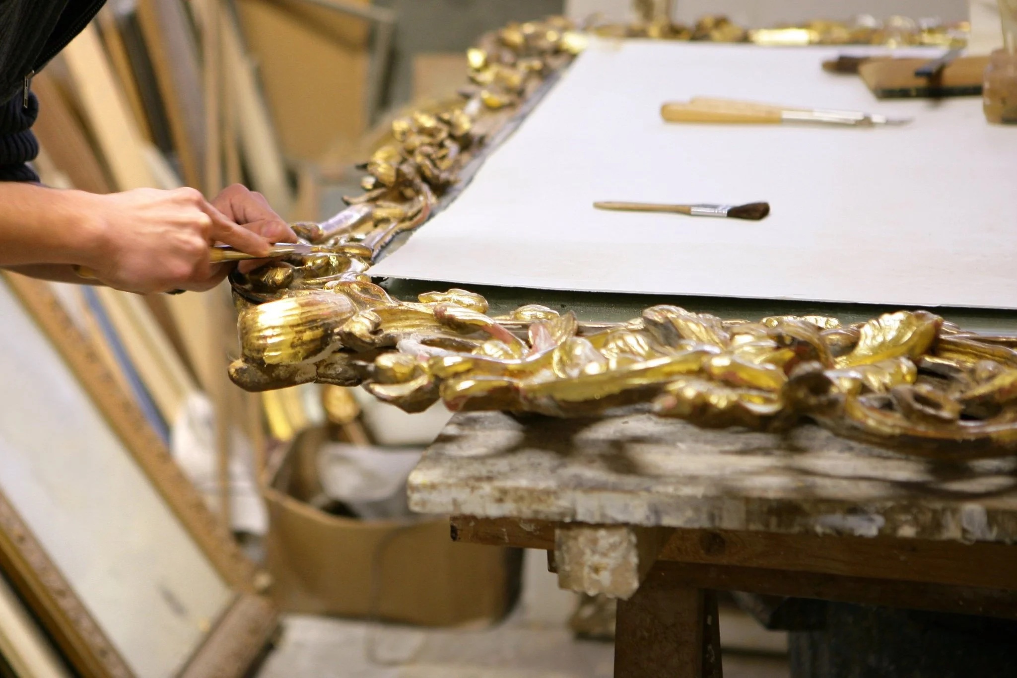 Hands applying gold leaf to an ornate antique frame during restoration in the Allart studio
